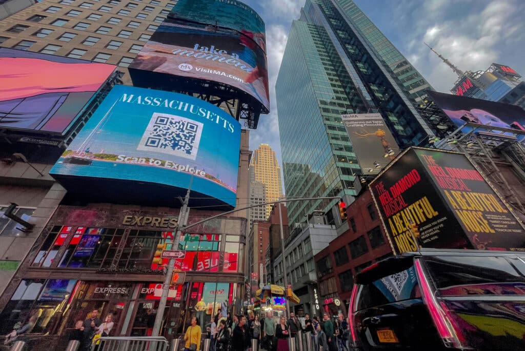 Colorful digital billboards in Times Square, New York City, including a Massachusetts tourism ad and a musical promotion.